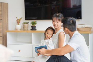 Fototapeta premium Padres asiáticos sentados en el piso de la sala en su hogar entreteniendo a su pequeña hija con un celular