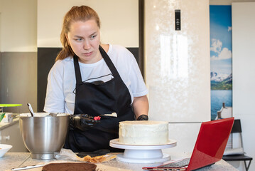 Pastry chef cook confectioner or baker in black gloves  makes a cake and using laptop. Food blogger recording video for followers, cooking channel. Homemade pastry, small home business 