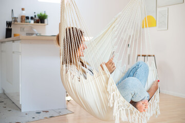 Fototapeta na wymiar Attractive happy young woman relaxing at home, lying in a hammock in living room, using smartphone, speaking on video chat, messenging with friend.