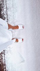 A girl in a white hotel coat and room white flip-flops in winter stands on snow-white snow. Dark feet in white slippers in the snow view from above