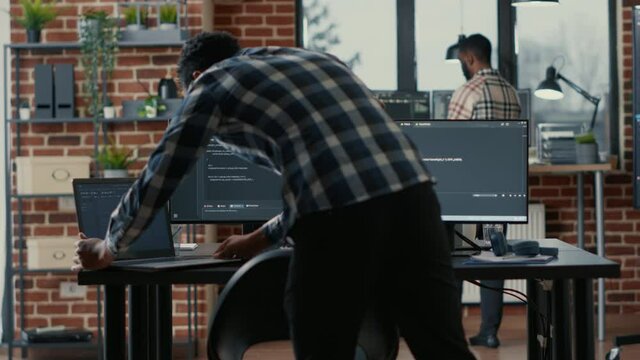 Programer coding on laptop sitting down at desk with computer screens parsing code in software agency. Software developer compiling algorithms with cloud programers working in the background.