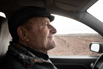 Old man smiling is driving by the countryside wearing a cap
