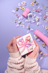 Woman hands hold cute gift box, high-colored Valentine day background with pink colored champagne bottle, spring blossom flowers, little heart decor, on violet very peri color background flatlay copy 