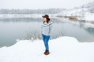 Fototapeta na wymiar Woman in a brown hat and sweater stands on shores of a snow-covered lake and forest