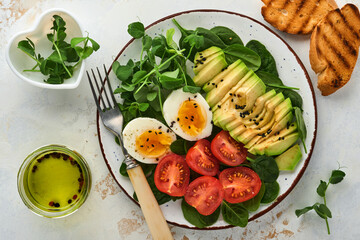 Avocado, cherry tomato, spinach and chicken egg, microgreens peas and black sesame seeds fresh salad in bowl on white stone table background. Healthy breakfast food concept. Top view.