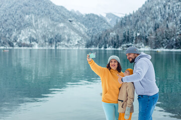 winter travel across Europe. view of the alpine lake with snow. portrait of happy family of travelers with child taking selfie on smartphone