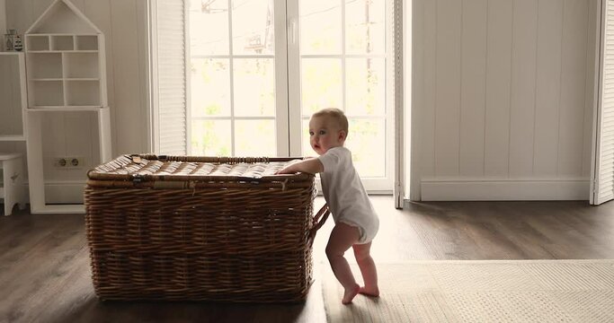 Cute active toddler makes first steps, growth and development, childhood concept. Adorable baby standing inside warm cozy sunny nursery room lean on wicker storage box, discover objects looks curious