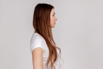 Side view portrait of woman looking to side copy space, feeling confident focused self-assured, expressing seriousness, wearing white T-shirt. Indoor studio shot isolated on gray background.