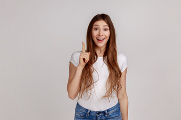 Portrait of inspired woman with long hair pointing finger up with genius idea, surprised by suddenly invented smart solution, wearing white T-shirt. Indoor studio shot isolated on gray background. - obrazy, fototapety, plakaty