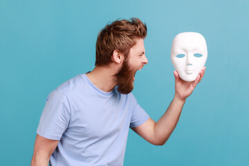 Portrait of bearded man screaming angrily, holding white mask in hands, hating his hidden...
