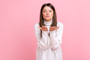 Romantic pretty woman sending air kisses, expressing love, flirts, looking at camera with pout lips, wearing white casual style sweater. Indoor studio shot isolated on pink background.