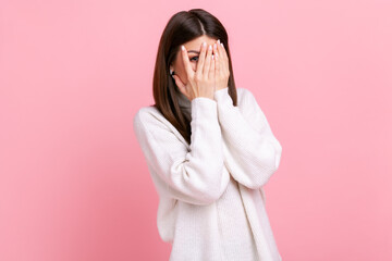 Curious woman looking through fingers, peeking with calm face, having suspicious, watching secrets, wearing white casual style sweater. Indoor studio shot isolated on pink background.