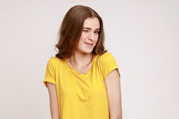 Obraz premium Closeup portrait of playful happy charming teenager girl with wavy hair in casual yellow T-shirt blinking eye, having cunning idea, flirting to camera. Indoor studio shot isolated on gray background.