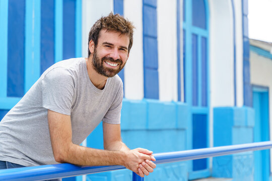 Cropped image of man lean on blue handrail with blue facade in the background. Horizontal view of latin american man on colorful wall with summer clothes. People and travel concept.