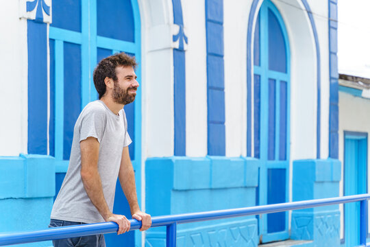 Cropped side view of man lean on blue handrail with blue facade in the background. Horizontal view of latin american man on colorful wall with summer clothes. People and travel concept.