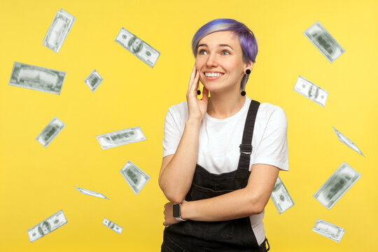 Portrait of happy beautiful hipster girl with violet short hair in denim overalls holding palm near face and dreaming about many money to have. isolated on yellow background, studio shot