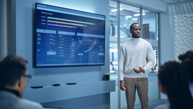 Office Conference Room Meeting Presentation: Black Businessman Talks, Uses Wall TV to Show Company Growth with Big Data Analysis, Graphs, Charts, Infographics. Multi-Ethnic e-Commerce Startup Workers