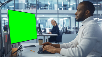 Diverse Modern Office: Handsome Black Businessman Using Desktop Computer with Green Screen Mock Up...