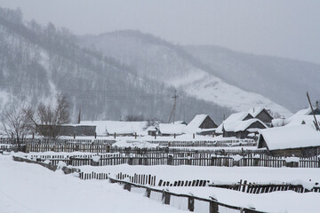 Snowfall in the village against the background of mountains. Houses, outbuildings, fences in the snow.
