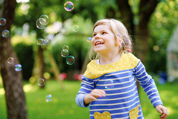 Happy little blonde preschool girl having fun with blowing soap bubble blower. Cute child playing...