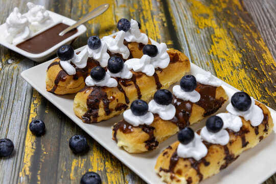 Delicious traditional French eclairs with vanilla cream, chocolate, cream and blueberries on a rustic wooden background.
