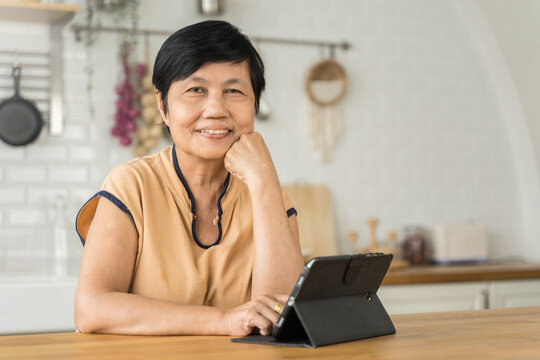 Smiling middle aged mature Asian woman looking at camera while using digital tablet. Portrait of beautiful senior female at home.