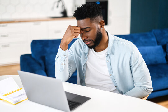 Tired frustrated black man sitting with eyes closed and using laptop at home, holding bridge of the nose, feels despair, African-American male student fail exams, has problems with remote work
