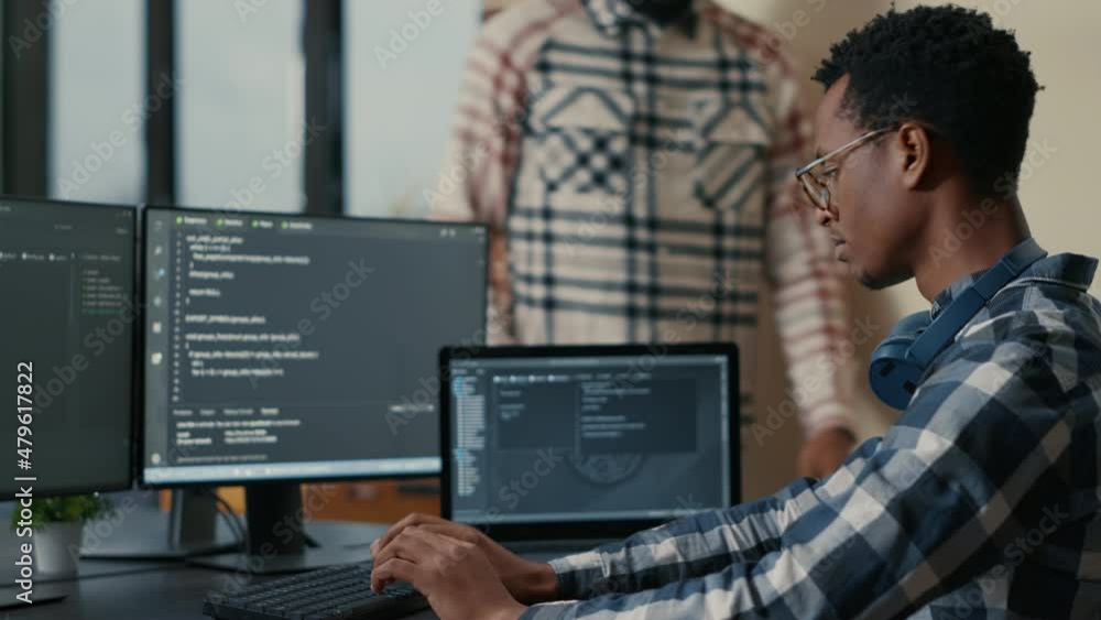Poster focused software developer writing code on laptop looking at multiple screens with programming langu