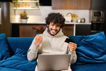 Smiling Indian man holding banking card and laptop, making purchases or ordering online. Mixed-race...