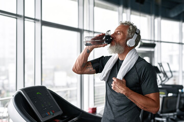 Mature athletic man takes care of health, drinks while exercising in gym - obrazy, fototapety, plakaty