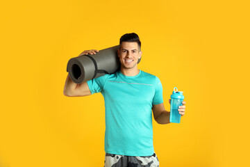 Handsome man with yoga mat and shaker on yellow background