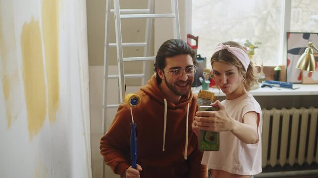 Young couple man and woman painting walls in yellow and taking photos selfie on smartphone while making overhaul. Husband and wife doing renovating together. Interior design, new apartment. 