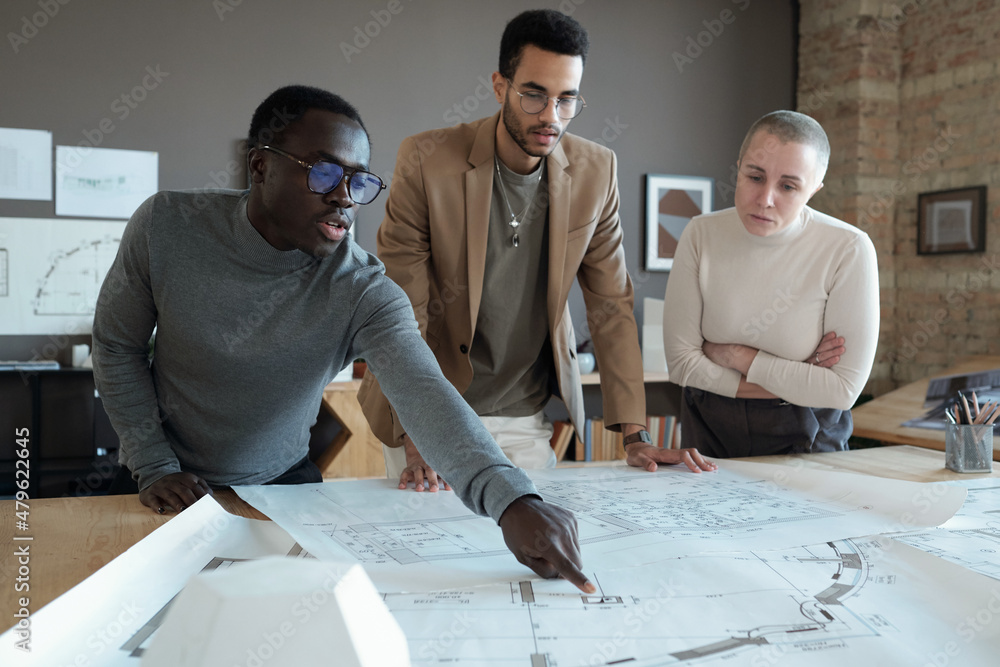 Poster Young confident architect pointing at blueprint while bending over table and explaining sketch to colleagues at meeting