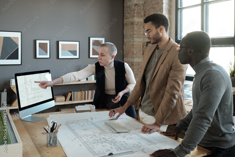 Poster mature businesswoman pointing at sketch on computer screen while explaining it to intercultural male