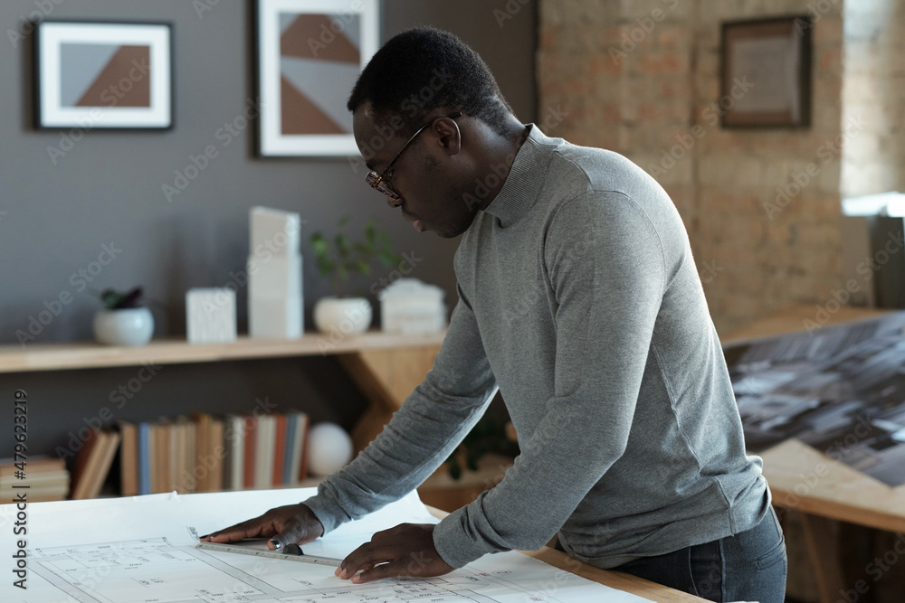 Poster Contemporary architect with ruler making sketch of new construction on blueprint while standing by table in office