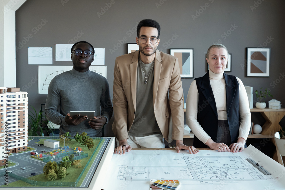 Poster Three contemporary professionals of various ethnicities standing by table with blueprints and house layout and looking at camera