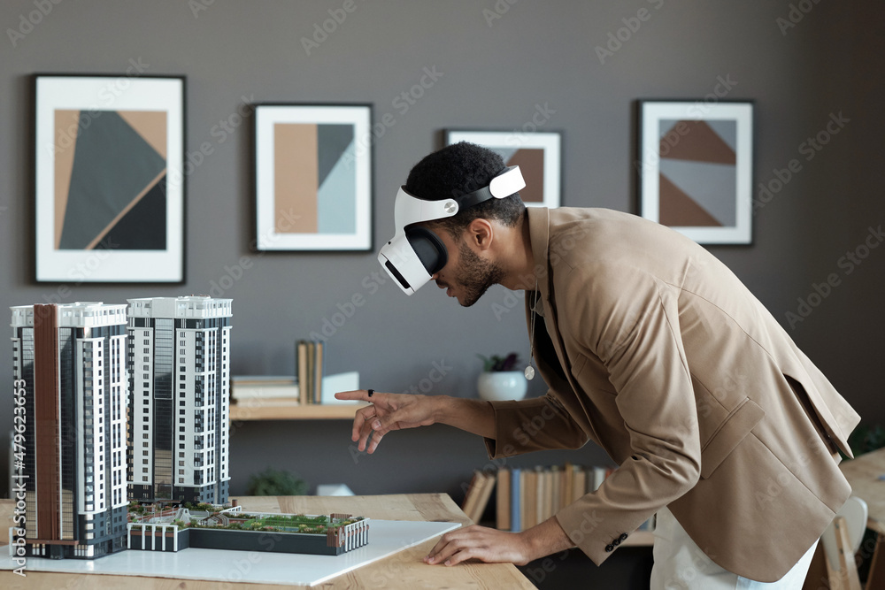 Poster young biracial businessman in vr headset bending over table and pointing at house layout while makin