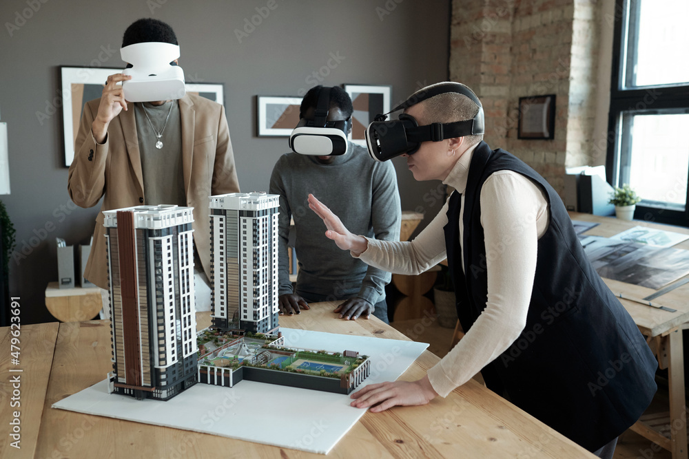 Poster Group of three architects in vr headsets standing by table with layout of new block of flats while one of them making presentation