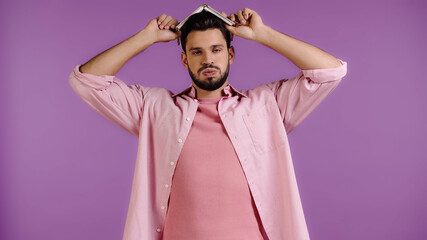 tired man puffing cheeks and holding book above head isolated on purple
