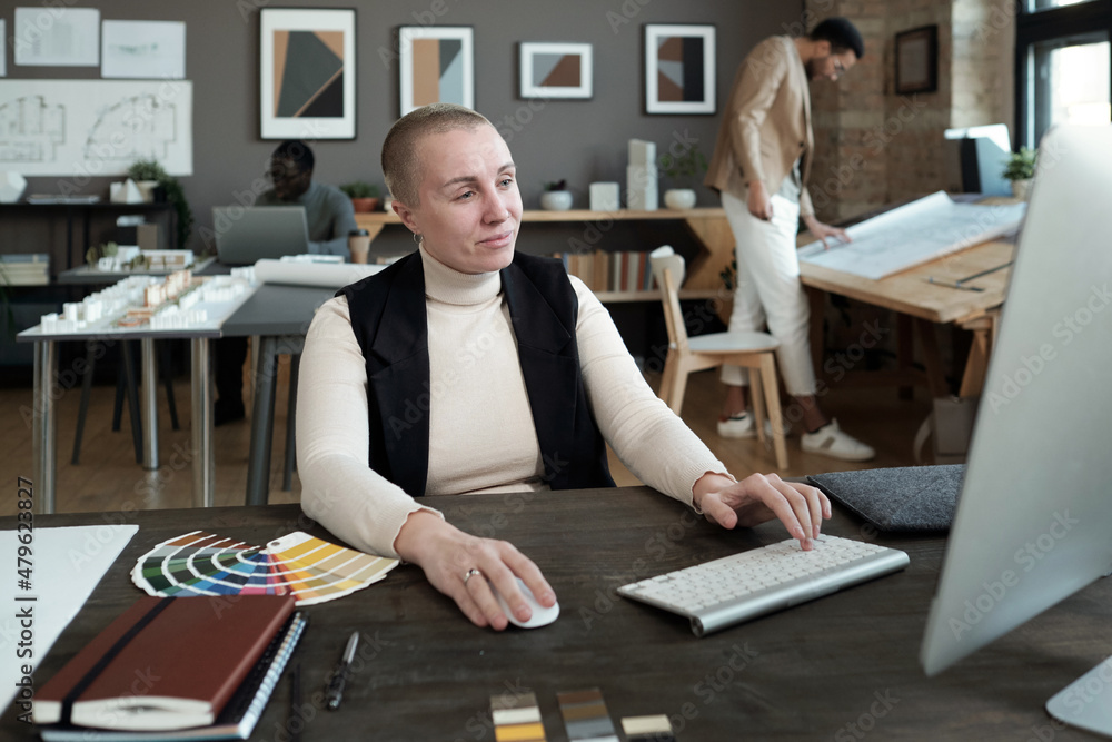 Poster mature female designer looking at computer screen while working by table against two intercultural m