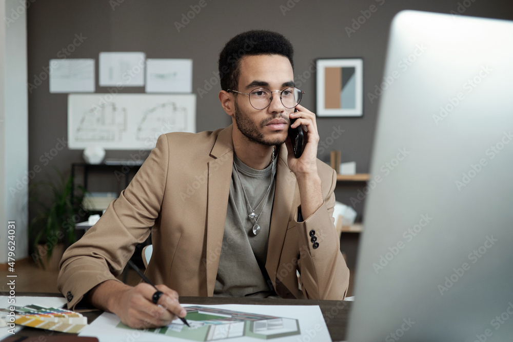 Poster Young serious designer with smartphone by ear looking at computer screen while consulting client and working over new project