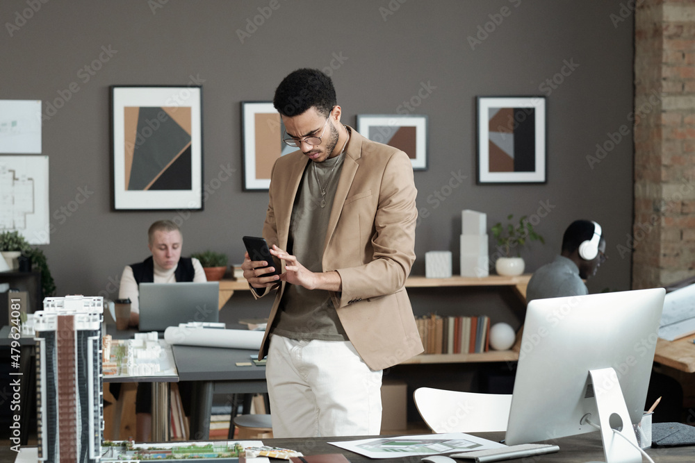 Poster Young biracial businessman scrolling in smartphone while standing by workplace with computer monitor and house layout