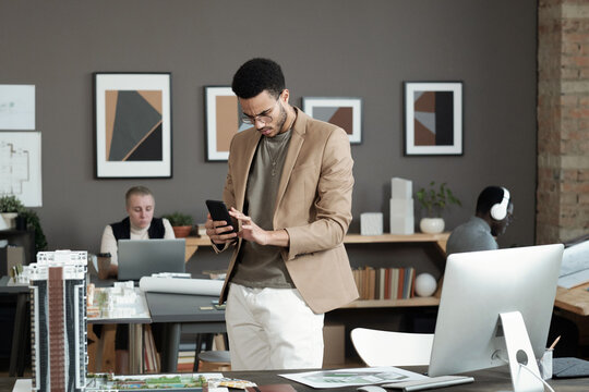 Young biracial businessman scrolling in smartphone while standing by workplace with computer monitor and house layout - Powered by Adobe