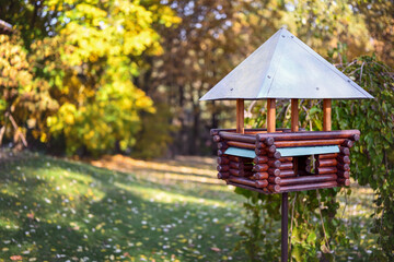 Red wooden bird feeder (birdhouse) with a metal roof in a city park. Sunny day, autumn. The feeder...