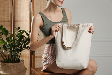 Happy young woman with blank eco friendly bag sitting on wooden stool indoors, closeup