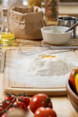Pile of flour with broken raw egg on wooden board surrounded by bowl, paper sack, fresh ripe tomatoes and bottle of olive oil