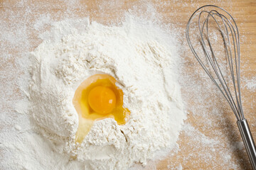 Flatlay of broken raw egg on top of pile of flour prepared for making homemade dough and whisk on wooden table