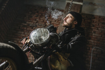 Fototapeta na wymiar Motorbiker in the black leather jacket smokes the cigarette near the the old motorbike concept.
