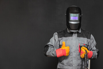 Welder man in the helmet and with welding terminals in the hand on the black wall background with copy space shows a thumbs up gesture.