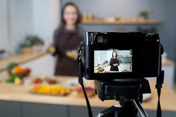 Video camera with happy young woman on screen holding bowl with homemade sauce during livestream in the kitchen